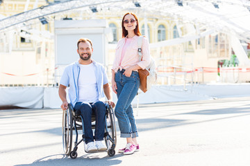 happy handsome boyfriend in wheelchair and girlfriend looking at camera on street