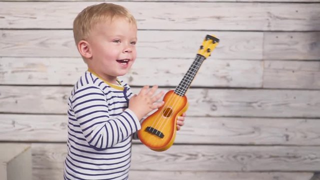happy one year old boy plays his guitar or ukulele and sings songs, sitting in the wooden room.