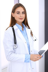 Doctor woman using tablet computer while standing straight near window in hospital. Happy physician at work. Medicine and health care concept