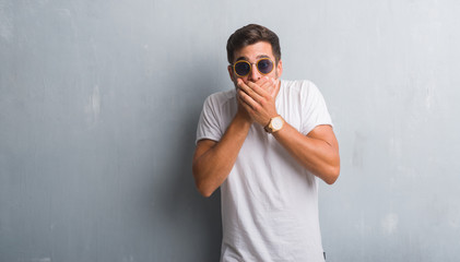 Handsome young man over grey grunge wall wearing sunglasses shocked covering mouth with hands for mistake. Secret concept.