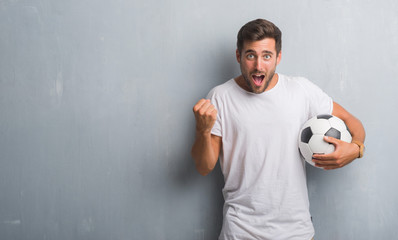 Handsome young man over grey grunge wall holding soccer football ball screaming proud and celebrating victory and success very excited, cheering emotion