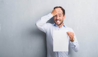 Handsome young man over grey grunge wall holding blank paper sheet contract stressed with hand on...