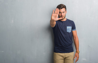 Handsome young man over grey grunge wall doing stop sing with palm of the hand. Warning expression with negative and serious gesture on the face.