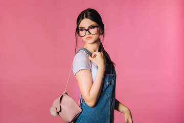 Fototapeta premium Girl in stylish clothes. Fashion. Trendy charming girl in colorful clothes, wearing black eyeglasses. Accessories. Backpack. Pink background. Fashionable studio of teenage style. Fashionable.