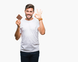 Young handsome man eating chocolate bar over isolated background doing ok sign with fingers, excellent symbol