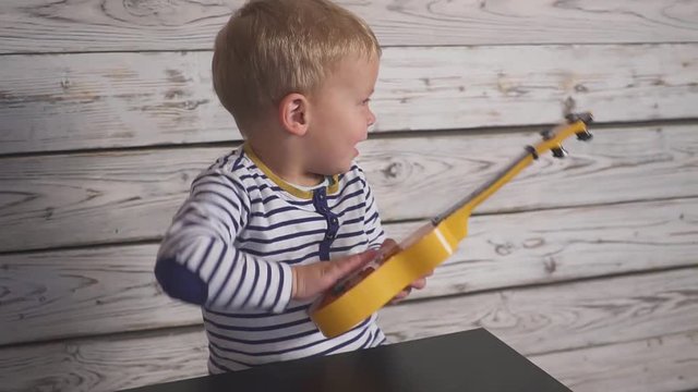 happy one year old boy plays his guitar or ukulele and sings songs, sitting in the wooden room.