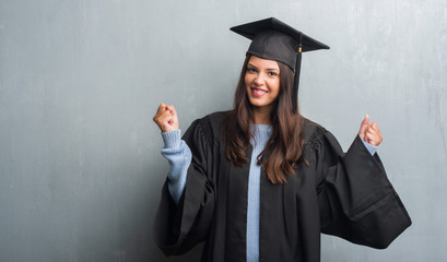 Young brunette woman over grunge grey wall wearing graduate uniform screaming proud and celebrating victory and success very excited, cheering emotion