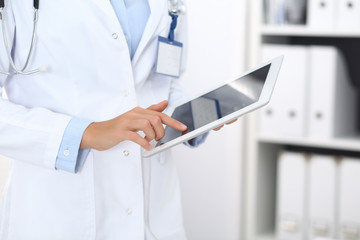 Doctor woman using tablet in clinic office. Unknown female physician at work while standing straight at hospital, close-up. Medicine and healthcare concept