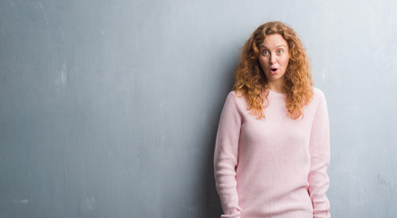 Young redhead woman over grey grunge wall wearing pink sweater afraid and shocked with surprise expression, fear and excited face.