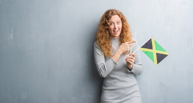 Young redhead woman over grey grunge wall holding flag of Jamaica very happy pointing with hand and finger