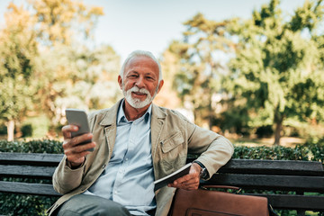Portrait of senior man using phone outdoors. Sitting on the bench and looking at camera.