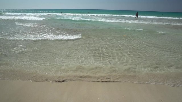 Clear skies and blue waters, view from the sandy beach as mild waves beautifully arrive to the shore, few people in ocean