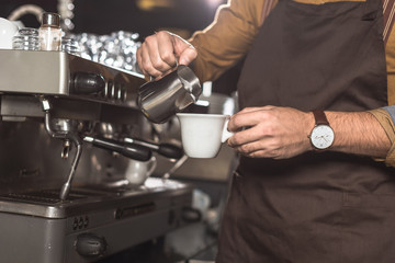 cropped shot of barista in apron pouring milk into coffee while preparing it in restaurant