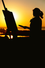 Fototapeta na wymiar silhouette of a woman painting a picture with paints on canvas on an easel, girl with paint brush and palette engaged in art on the nature in a field at sunset