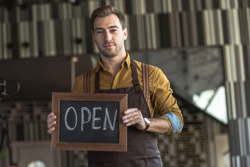 attractive young waiter holding board with open inscription in cafe