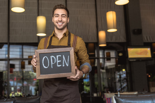 attractive smiling waiter holding chalkboard with open inscription in cafe