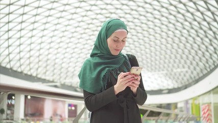 Pretty muslim woman in a hijab stands in a shopping center and uses the phone