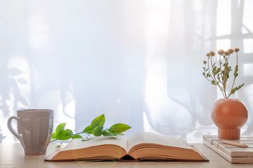 Hipster wooden desk with a vase of dried plants, an open thick book and a gray cup with coffee set under the window. Copy space