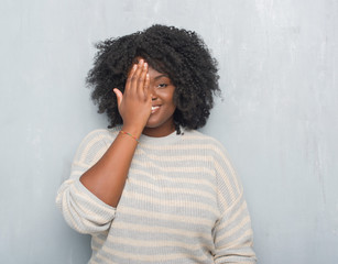 Young african american plus size woman over grey grunge wall wearing a sweater covering one eye with hand with confident smile on face and surprise emotion.