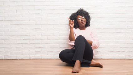 Young african american woman sitting on the floor at home pointing finger up with successful idea....