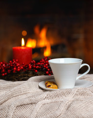 Cup of hot drink with cookie berries and red candle in red Christmas decoration on cozy knitted plaid in front of fireplace. Christmas New Year concept. Cozy relaxed magical atmosphere home interior.