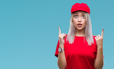 Young blonde woman wearing red hat over isolated background amazed and surprised looking up and pointing with fingers and raised arms.