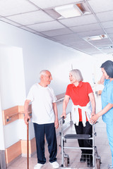 Elderly couple with stick and walker moving in hospital hallway assisted by nurse