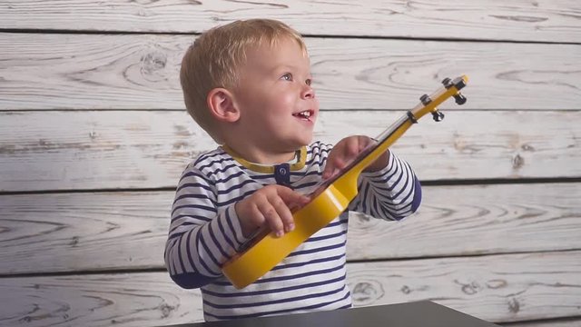 happy one year old boy plays his guitar or ukulele and sings songs, sitting in the wooden room.