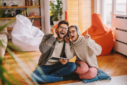 Happy couple listening to favorite song while sitting on the floor. Man holding tablet and woman having headphones on ears. Living room interior.