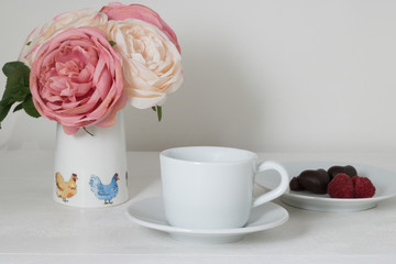 Obraz na płótnie Canvas White cup, two dark heart-shaped chocolates, fresh raspberry berries, pastel-colored flowers in a vase are on a white wooden table against a white wall.