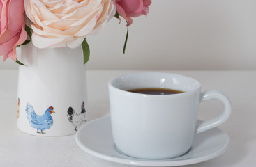 Close-up: white cup of tea or black coffee and a vase with bouquet of flowers and pattern of cute chicken are on a white wooden table. 