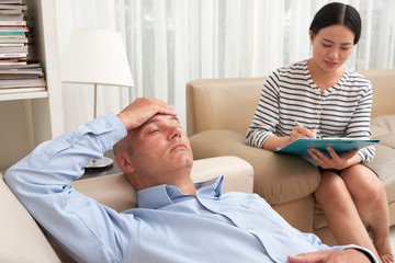 Fototapeta premium Mature man at the psychotherapist, lying on couch, discussing with psychotherapist his problems, practitioner making some notes