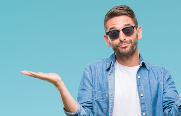 Young handsome man wearing sunglasses over isolated background clueless and confused expression with arms and hands raised. Doubt concept.