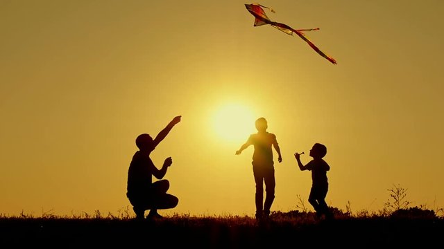 A happy father with two children playing in the open air running a flying kite. Silhouette of unrecognizable people at sunset.