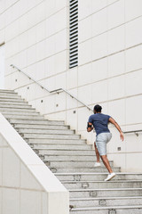 Rear view of unrecognizable sportsman with smartphone on shoulder running up concrete steps while having morning workout