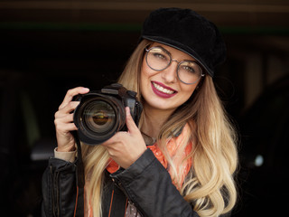 Portrait of a beautiful stylish cheerful young blonde photographer in glasses and a hat with makeup holding a black camera in a cookie against a dark background