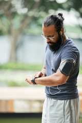 Muscular Indian man with long beard checking time on watch after running while standing outdoors on blurred background