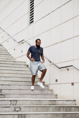 Muscular bearded tattooed Indian man in sportswear listening to music and running down concrete stairs outdoors