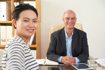 Portrait of confident smiling businessman sitting at his workplace with young woman, she came at meeting