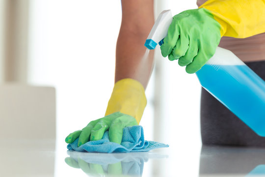 Young woman in protective gloves wiping dust using a spray and a cloth while cleaning her house