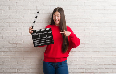 Young Chinese woman over white brick wall holding clapboard very happy pointing with hand and finger