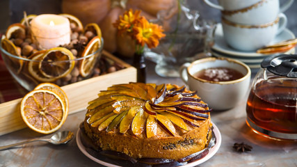 Plum cake decorated with sliced plum among the autumn decor - pumpkin, candle, oranges and yellow chrysanthemum. Rustic style. Homely cozy still life
