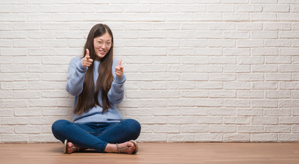 Young Chinese woman sitting on the floor over brick wall pointing fingers to camera with happy and funny face. Good energy and vibes.