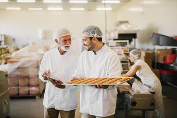 Fototapeta na wymiar Picture of two employees in sterile clothes in food factory smiling and talking. Younger man is holding tray full of fresh cookies while the older is holding tablet and checking production line.