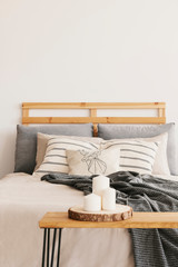 Three candles on the wooden table next to cozy bed with patterned pillows and grey blanket, real photo with copy space on the wall