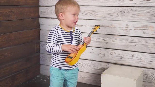 happy one year old boy plays his guitar or ukulele and sings songs, sitting in the wooden room.