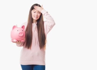 Young Chinese woman over isolated background holding piggy bank stressed with hand on head, shocked with shame and surprise face, angry and frustrated. Fear and upset for mistake.