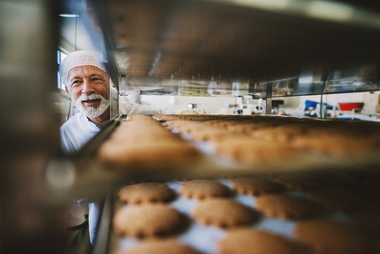 Picture of professional mature male baker man in white work uniform. Standing in front of the shelves full with fresh baked cookies.