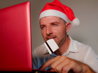 young happy and attractive man in Santa Klaus hat using credit card and laptop computer for buying...