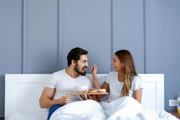 Happy young couple having breakfast in their bed. Beautiful girl is feeding her handsome boyfriend.